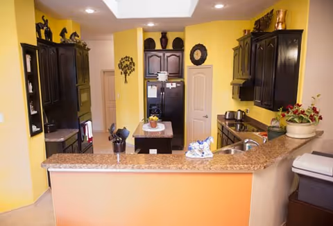 A kitchen with yellow walls and dark wood cabinets. There is a granite countertop with a sink and a small island in the center. The kitchen includes a black refrigerator, a stove, a toaster, and decorative items on top of the cabinets and island. The lighting is recessed in the ceiling.
