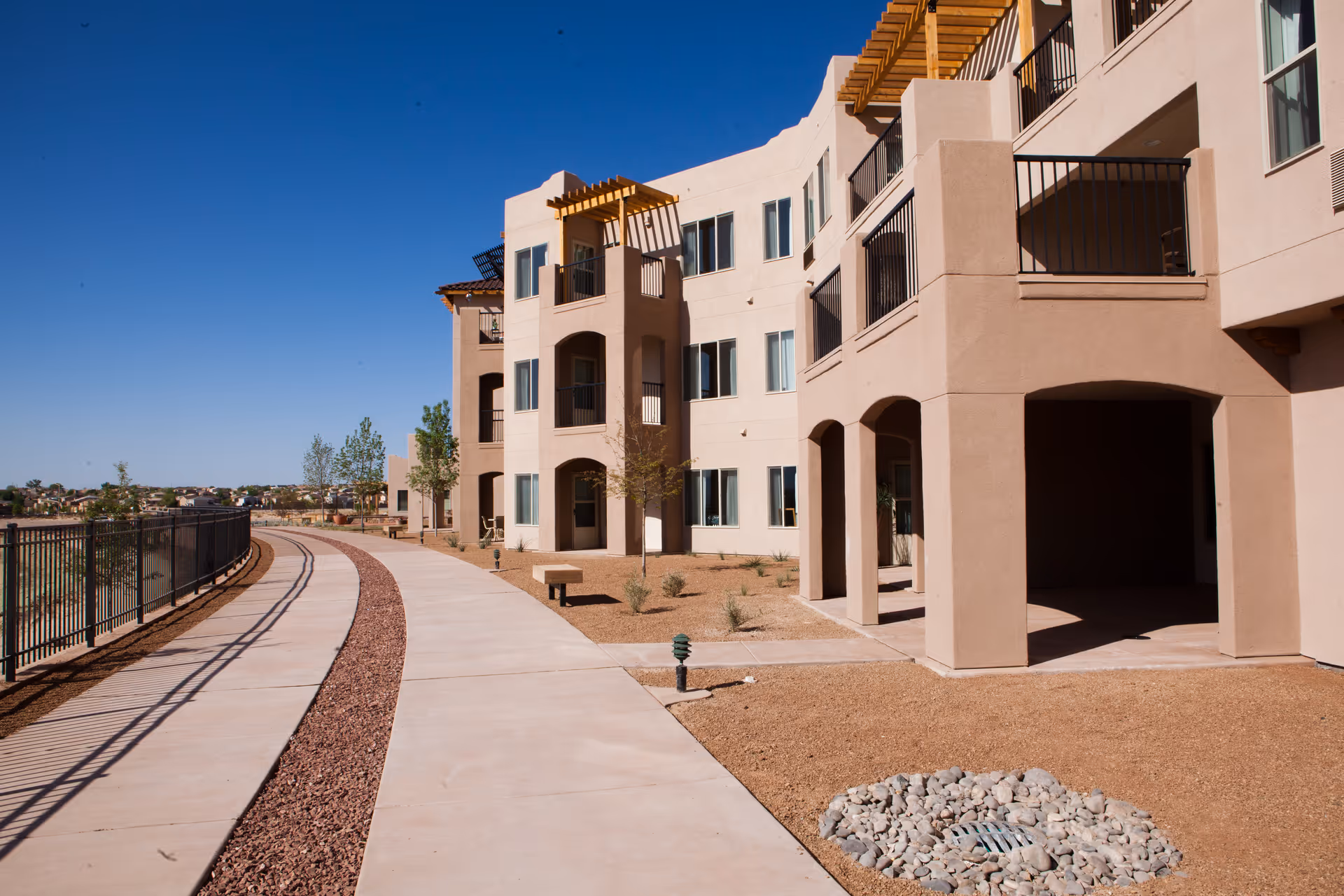 Exterior view of a multi-story beige retirement building with balconies and a curved walkway under a clear blue sky.
