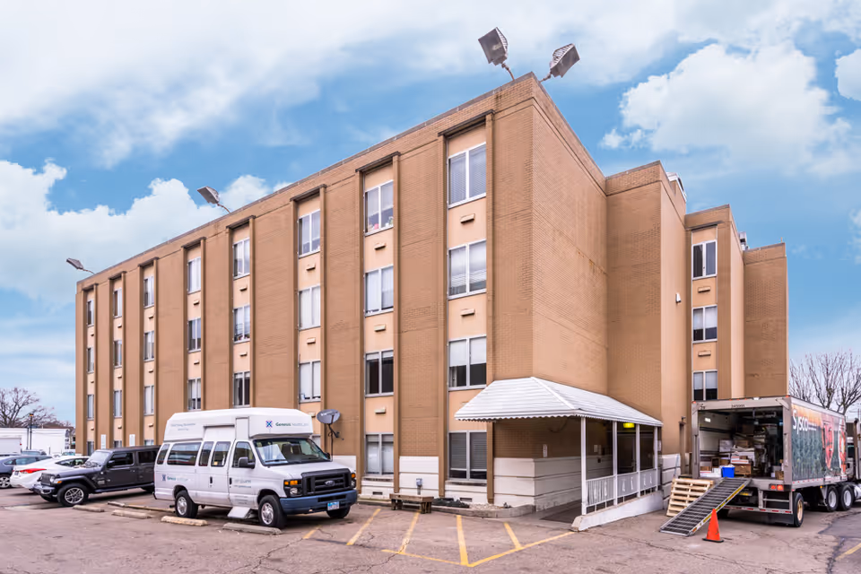 Brick multi-story nursing facility building with a covered entrance, parked vans and a delivery truck outside under a partly cloudy sky.