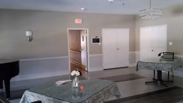 Communal dining/activity room with tables draped in green tablecloths, floral centerpieces, a piano at left, and doors on a beige wall.