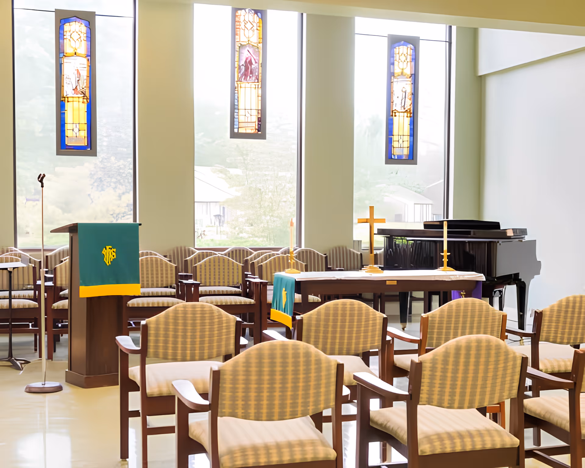 Chapel interior with rows of cushioned chairs facing a lectern, altar with a cross and piano beneath tall stained-glass windows.