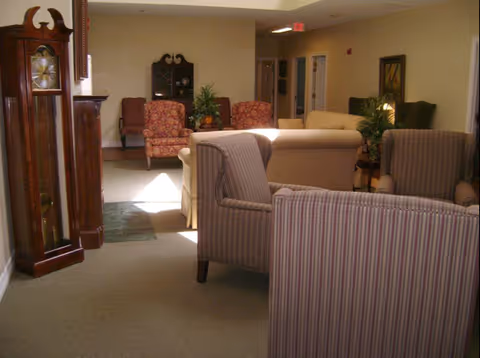 A cozy lounge area with upholstered sofas and armchairs, a grandfather clock on the left, potted plants, and a hallway in the background.