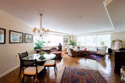 Bright senior living lounge with sofas, chairs, coffee tables, chandeliers, and large windows.