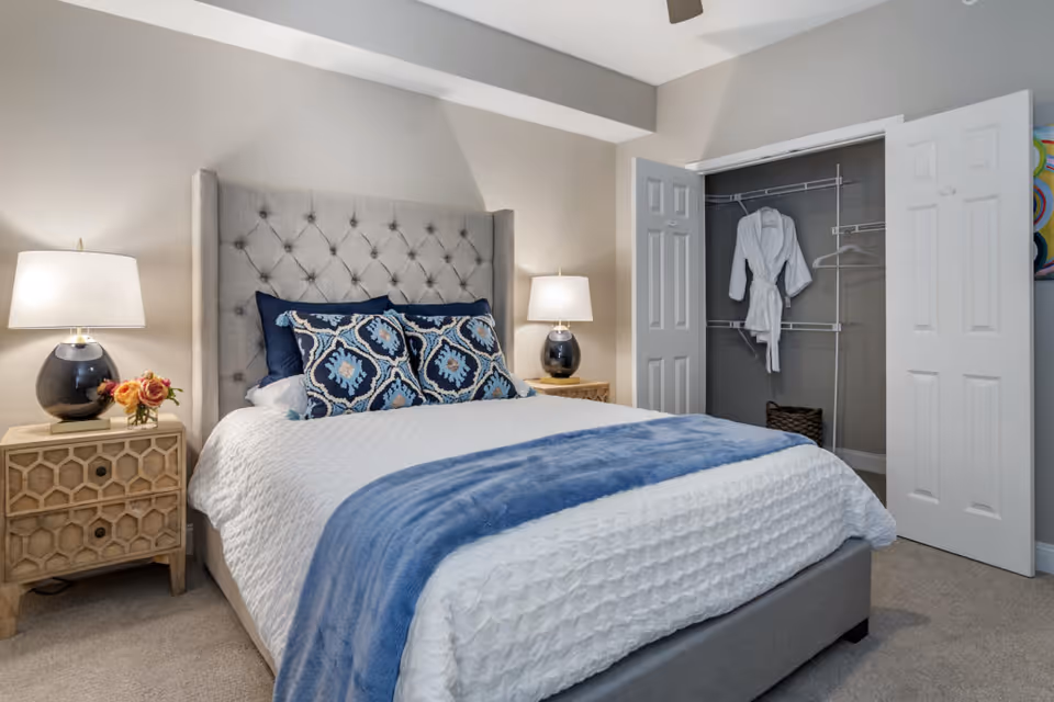 Well-lit bedroom with a tufted headboard, blue-and-white bedding, two bedside tables with lamps, and an open closet holding a white robe.