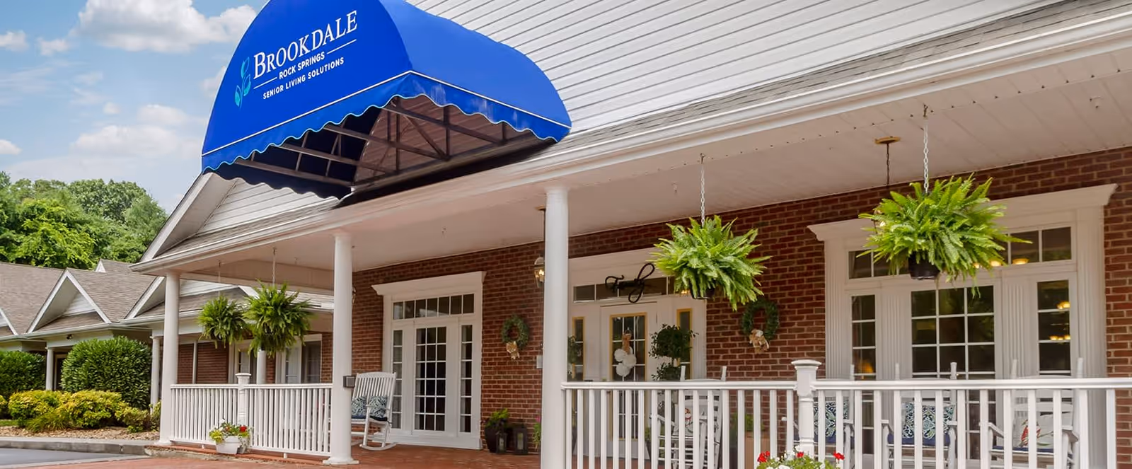 Exterior view of Brookdale Rock Springs senior living facility showing a brick building with white columns and railings, a blue awning with the facility name, hanging green plants, and a porch with rocking chairs.