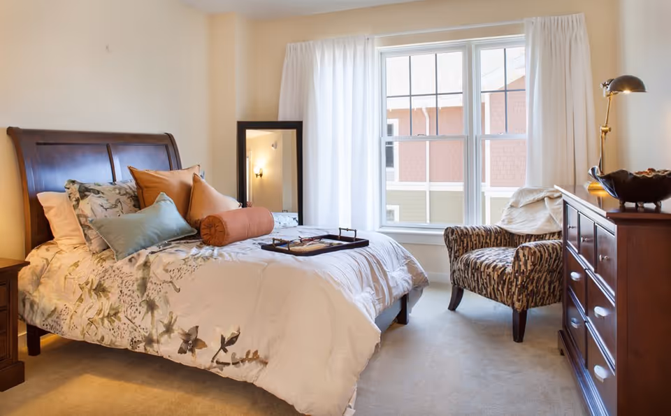 Bright furnished bedroom featuring a wooden bed with patterned bedding, decorative pillows, an armchair by a large window, and a wooden dresser with a lamp.