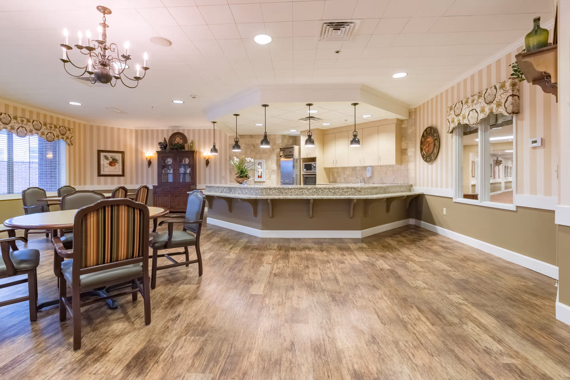 A spacious dining area with round tables and chairs, a chandelier hanging from the ceiling, and a kitchen counter with pendant lights. The room has wood flooring, striped wallpaper, and windows with decorative valances.
