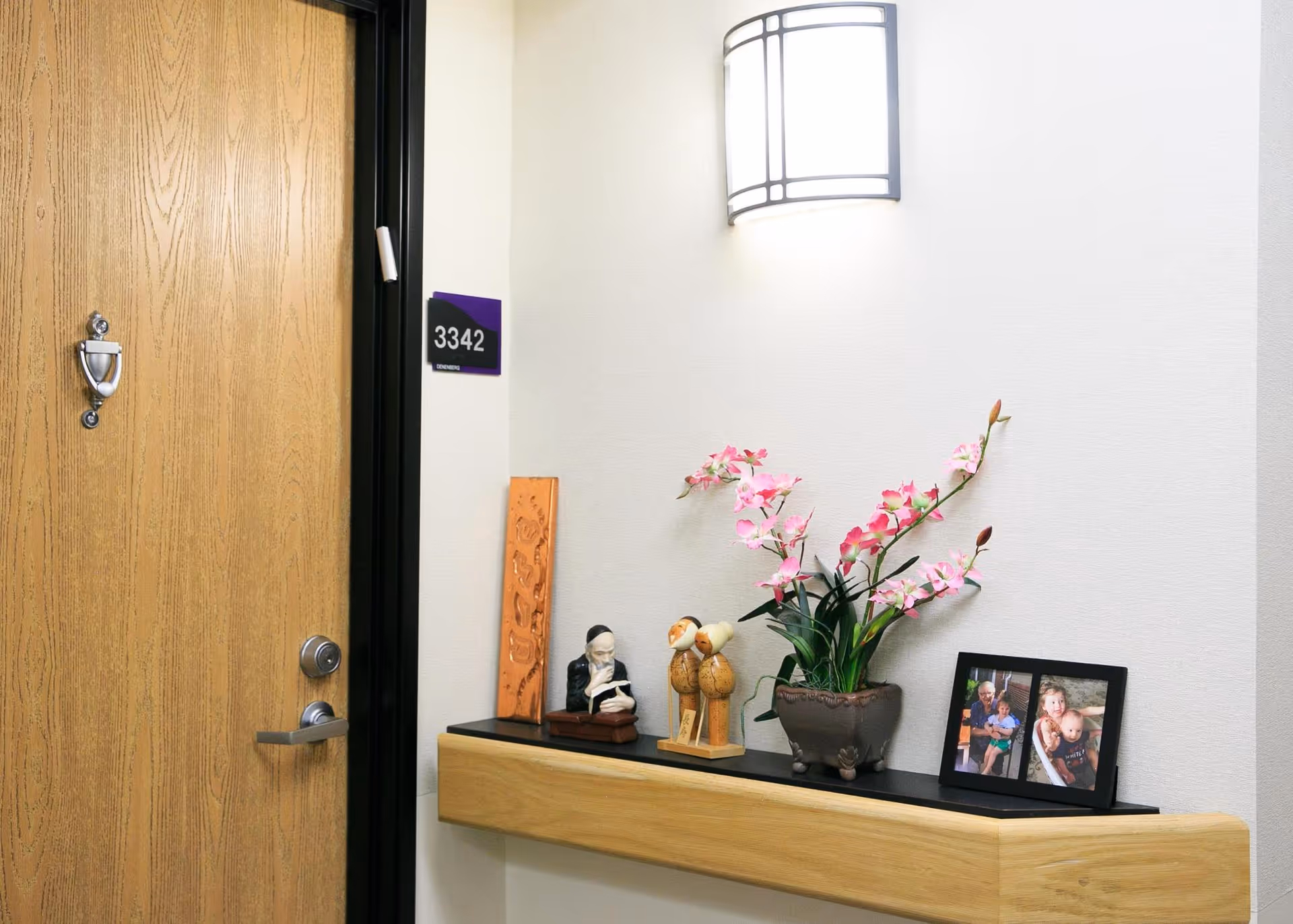 Hallway entry with a wooden door marked 3342 and a wall shelf holding flowers, figurines, and a framed photo under a wall sconce.