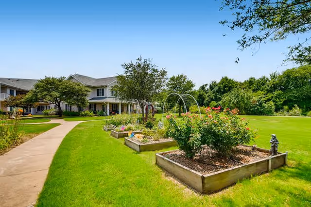 A well-maintained garden area with raised flower beds containing various plants and flowers, a concrete pathway curving through a green lawn, and a two-story building in the background under a clear blue sky.