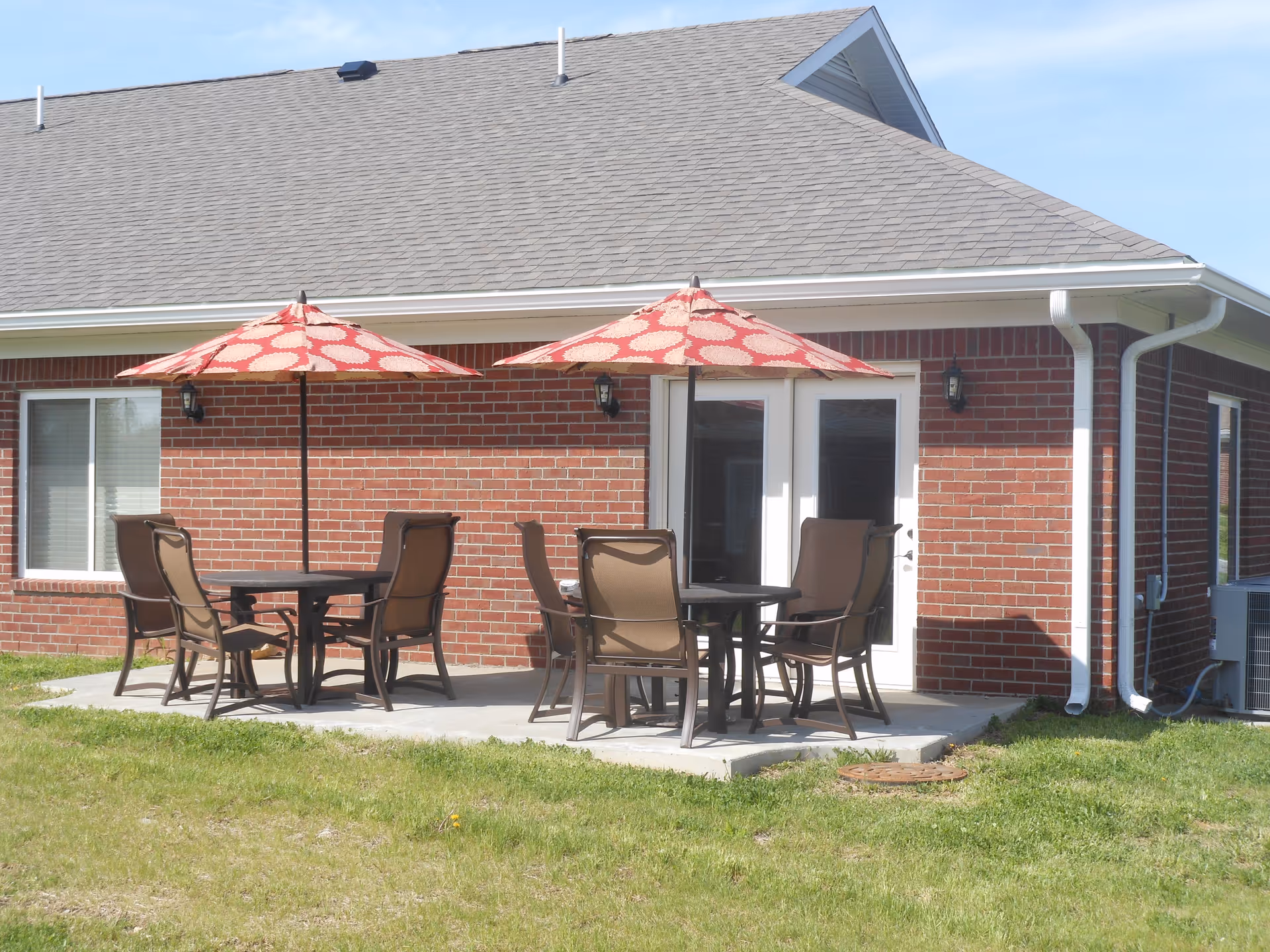 Brick building exterior with two patio tables, chairs, and red patterned umbrellas on a concrete patio.
