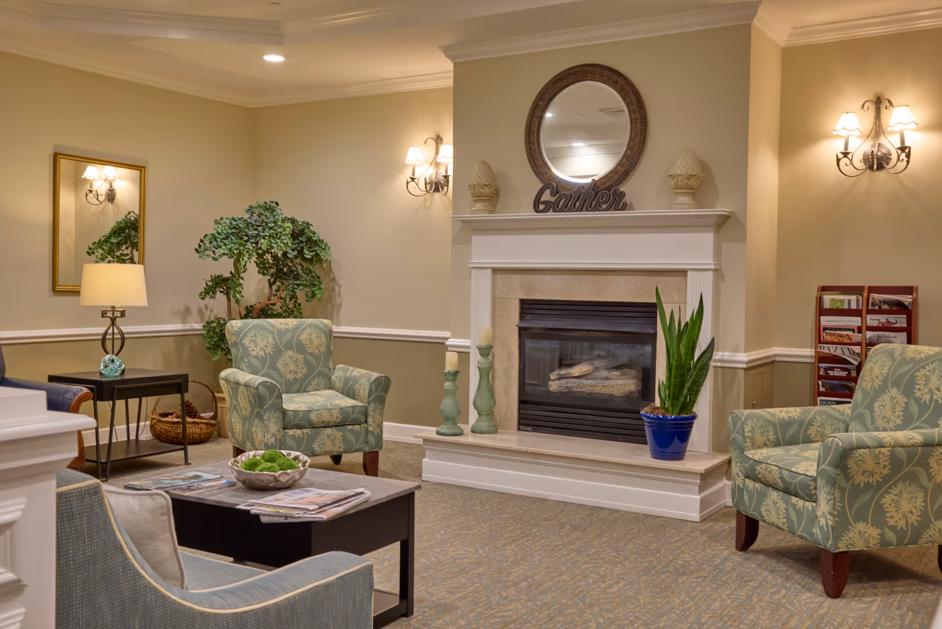 A cozy living room area with patterned armchairs, a coffee table with magazines and a decorative bowl, a fireplace with a round mirror and decorative items on the mantel, a potted plant, wall sconces, a side table with a lamp, and a magazine rack in the corner.