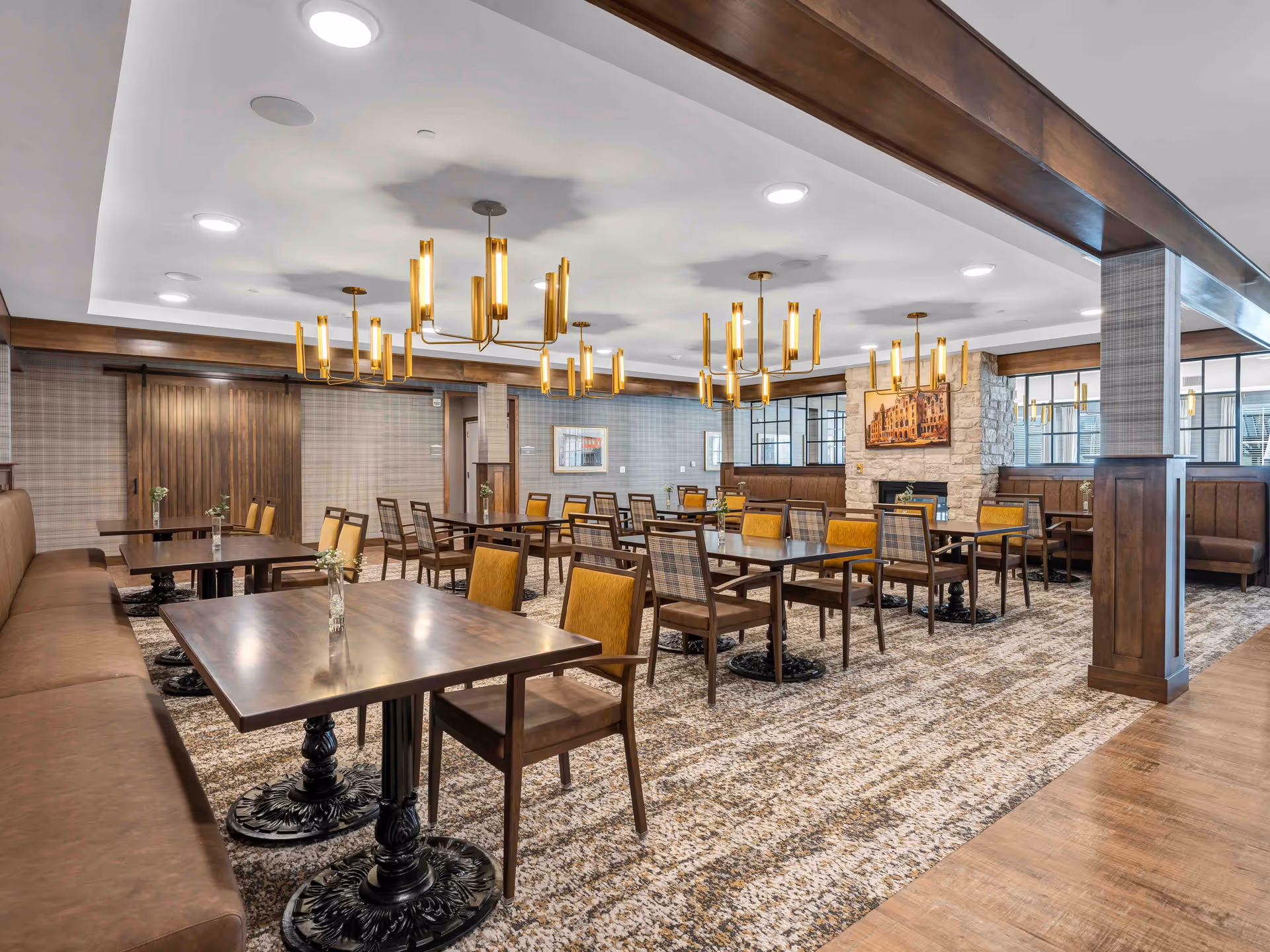 A spacious dining room in a senior living facility featuring multiple wooden tables with chairs, brown cushioned bench seating along the left wall, and modern gold chandeliers hanging from the ceiling. The room has a patterned carpet, wood paneling, and a stone fireplace with a framed picture above it.