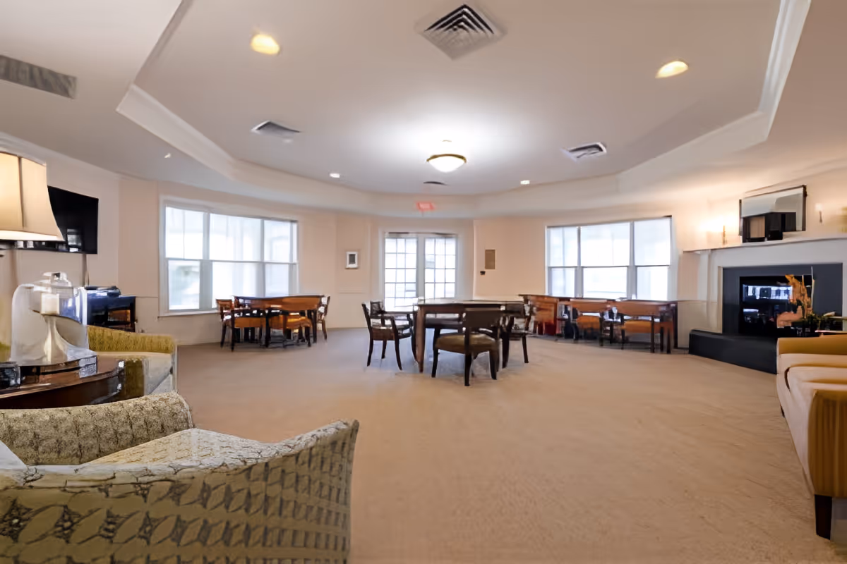 A spacious, well-lit common area with multiple seating arrangements including armchairs, tables, and chairs. Large windows allow natural light to fill the room. There is a fireplace on the right side and a television mounted on the left wall. The room has a beige carpet and a white ceiling with recessed lighting.
