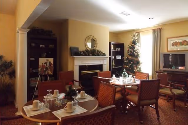 A cozy dining area decorated for the holidays with a Christmas tree, a fireplace with a mirror above it, and tables set with cups, glasses, and napkins. The room has warm lighting, red cushioned chairs, and bookshelves in the background.