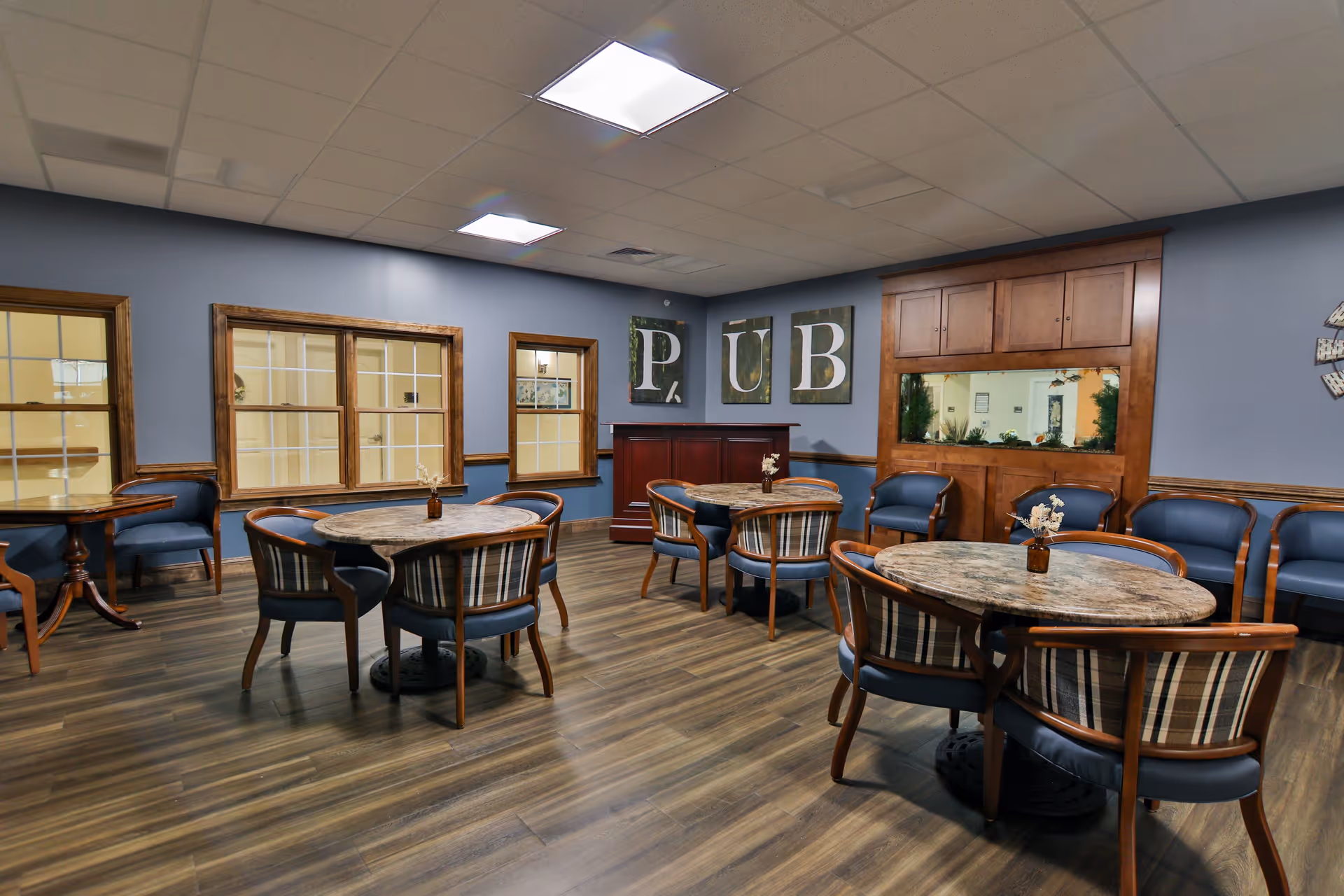 A cozy senior living common area with round tables and chairs arranged for socializing. The walls are painted blue with wooden trim, and there are three large framed letters spelling 'PUB' on one wall. A built-in wooden cabinet with a fish tank is also visible, along with a row of blue cushioned chairs against the wall.