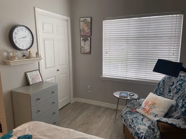 A cozy room with a window covered by white blinds, a blue patterned armchair with a pillow that says 'Hello Sunshine', a small round side table with a book and glasses, a gray chest of drawers with a framed picture on top, a wall clock above a small shelf with decorative items, and two framed pictures on the wall next to a closed white door.