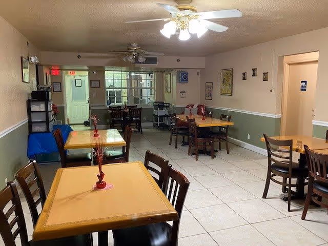 Interior view of a dining room in an assisted living facility with several wooden tables and chairs arranged neatly. Each table has a small red centerpiece. The room has tiled floors, beige walls with a green lower half, ceiling fans with lights, and framed pictures on the walls. There is a doorway leading to another room and an exit door visible in the background.