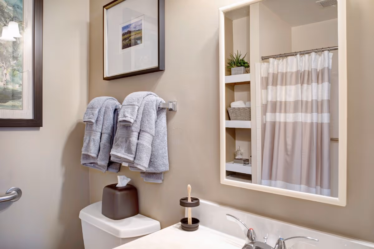 Bathroom with folded gray towels on a wall rack, a toilet and tissue box, sink and mirror reflecting a shower with a striped curtain.