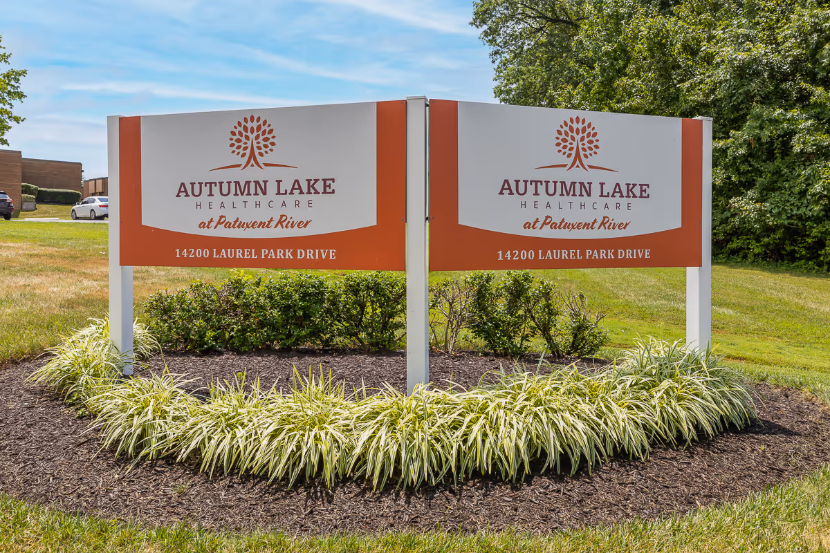 Outdoor view of a double-sided sign for Autumn Lake Healthcare at Patuxent River, located at 14200 Laurel Park Drive, surrounded by landscaping with green grass, bushes, and plants under a blue sky.
