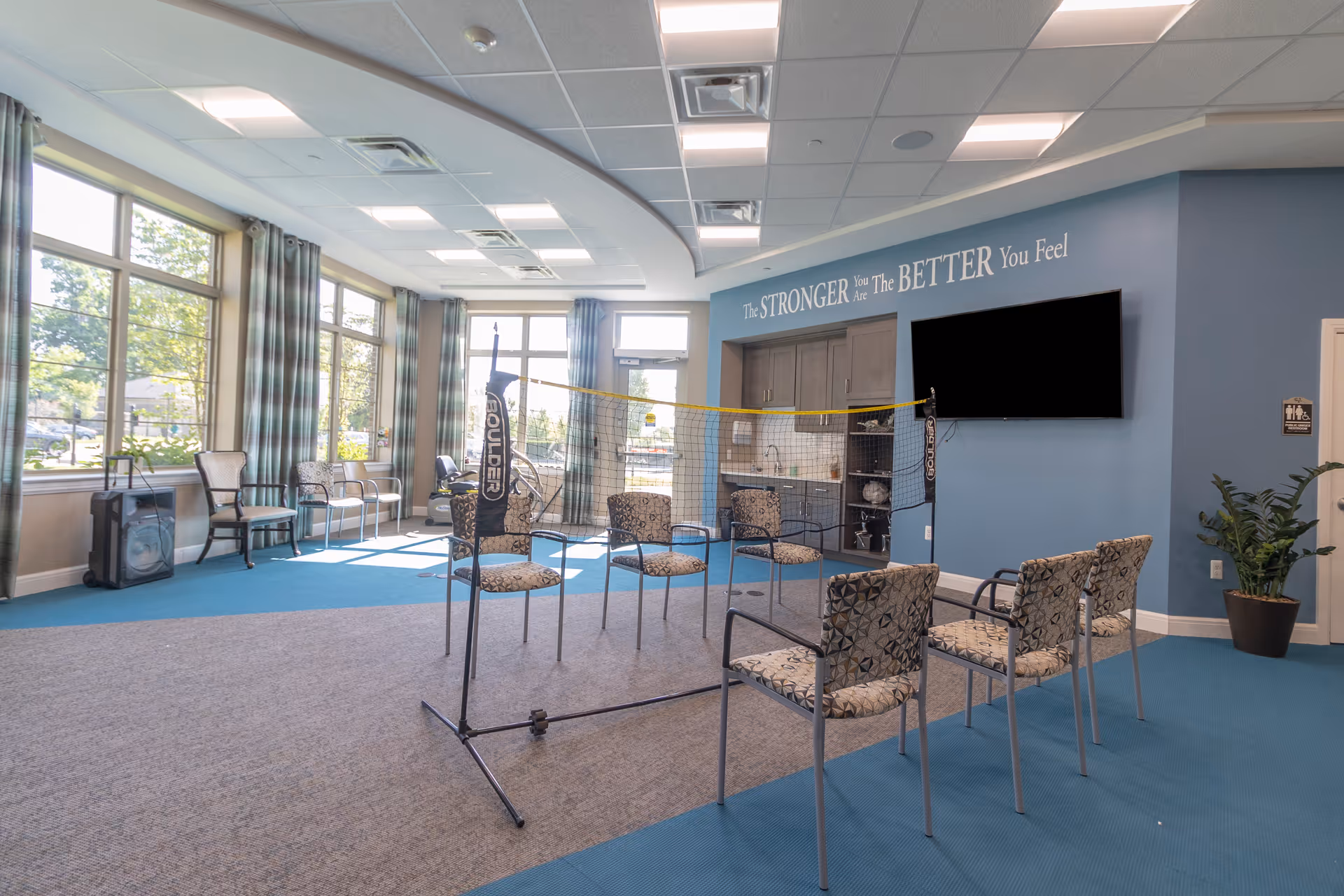 Bright communal activity room with chairs arranged around a portable net, large windows, a wall-mounted TV, and a small kitchenette area.
