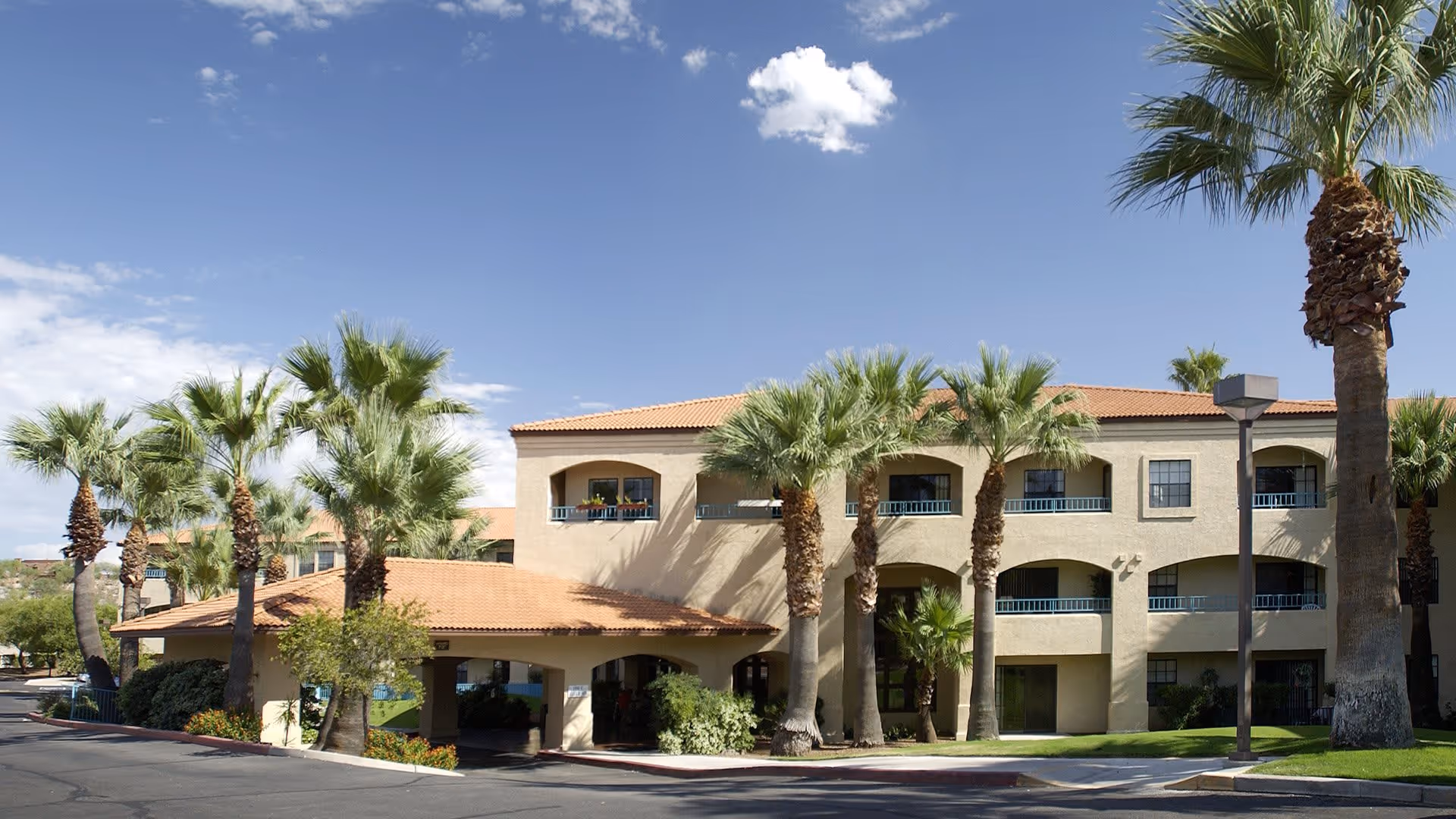 Exterior view of a senior living facility building with beige walls and a red-tiled roof, surrounded by palm trees under a clear blue sky with a few clouds.