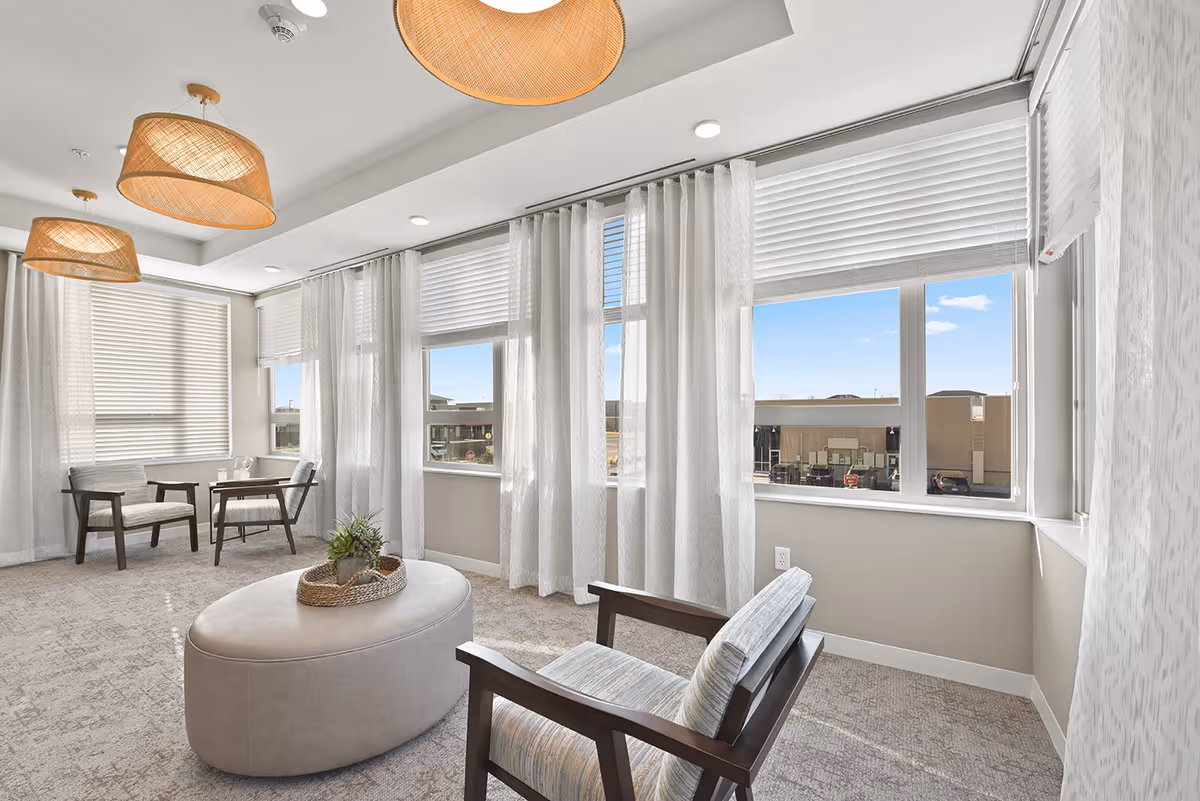 Bright lounge with chairs, an ottoman, a small table and large windows with sheer curtains.