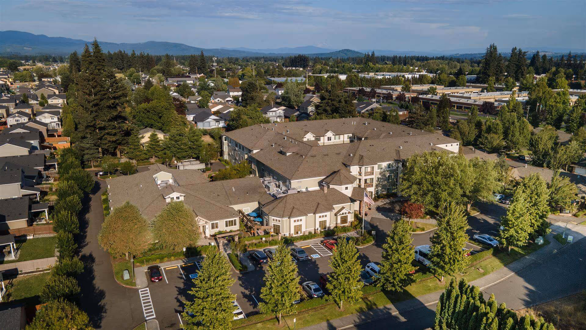 Aerial view of Brookdale Vancouver Orchards senior living facility surrounded by trees and residential houses, with a parking lot in front and mountains visible in the background under a partly cloudy sky.