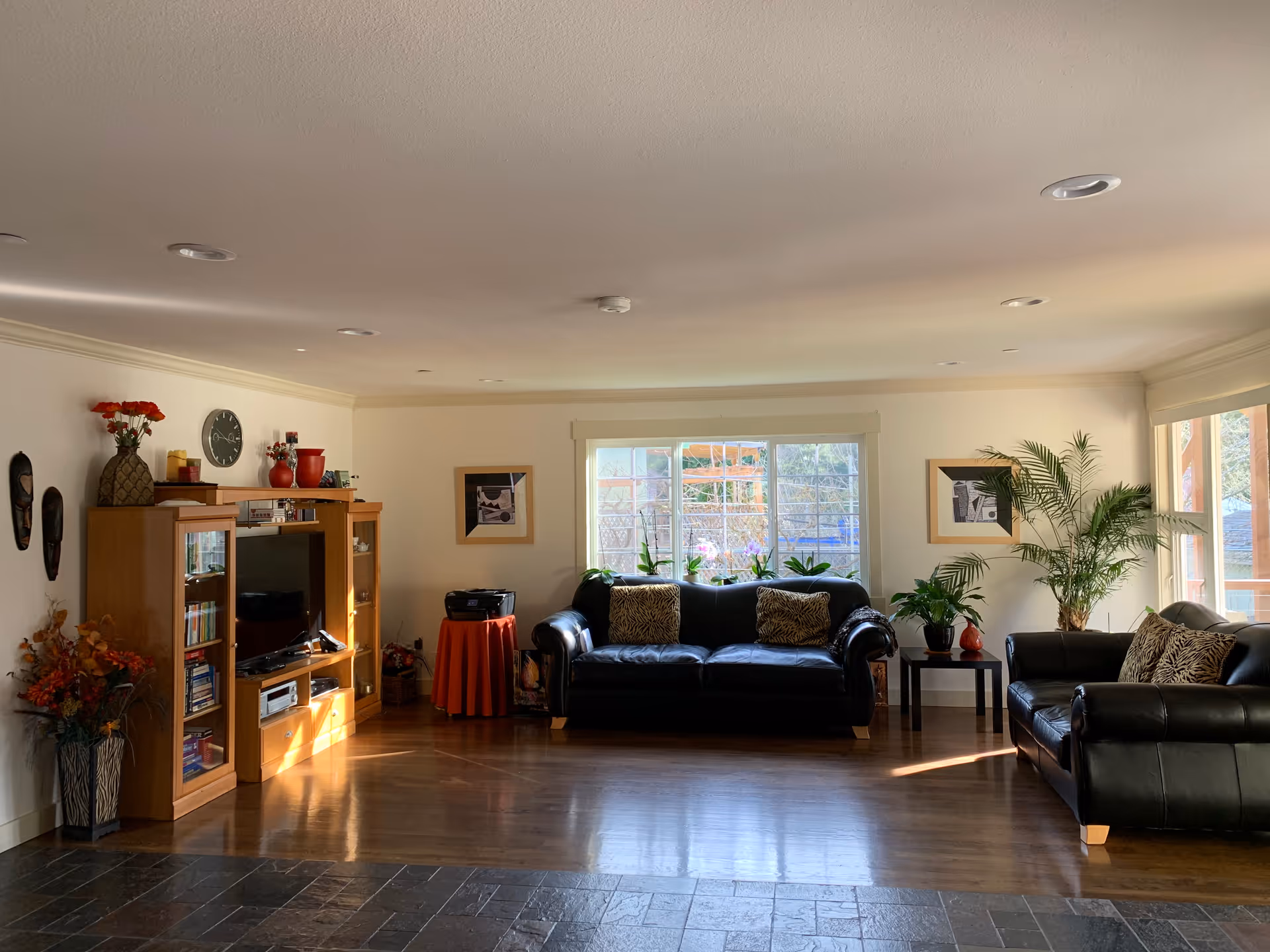 A bright and spacious living room with two black leather sofas adorned with patterned cushions. There is a wooden entertainment center with a TV, books, and decorative items on the left side. The room has large windows letting in natural light, with plants and framed pictures adding to the cozy atmosphere. The floor is a combination of dark tiles and wood.