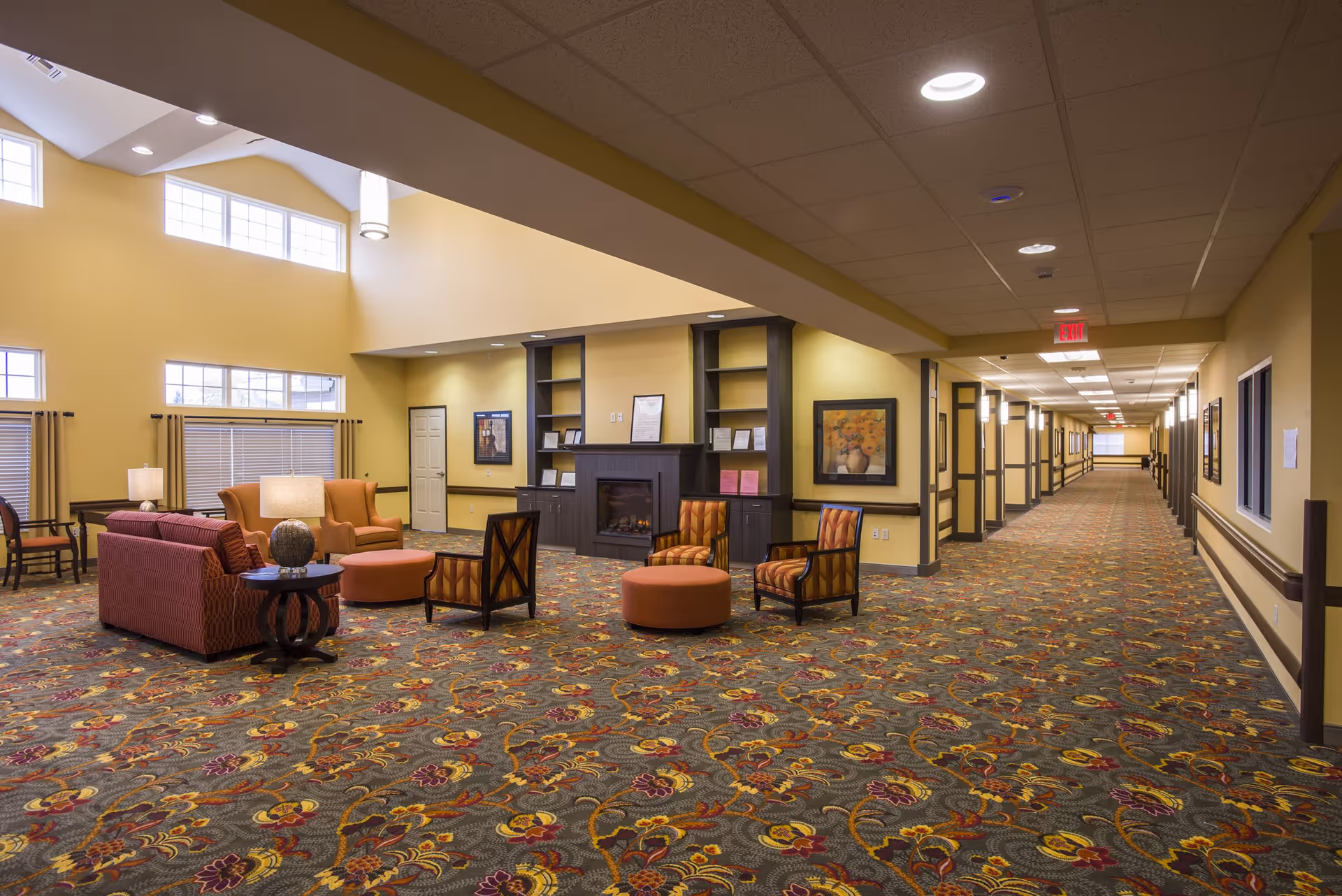 A spacious and well-lit common area in a senior living facility with patterned carpet, several upholstered chairs and sofas arranged around a fireplace, large windows with blinds, and a long hallway extending to the right with framed artwork on the walls.