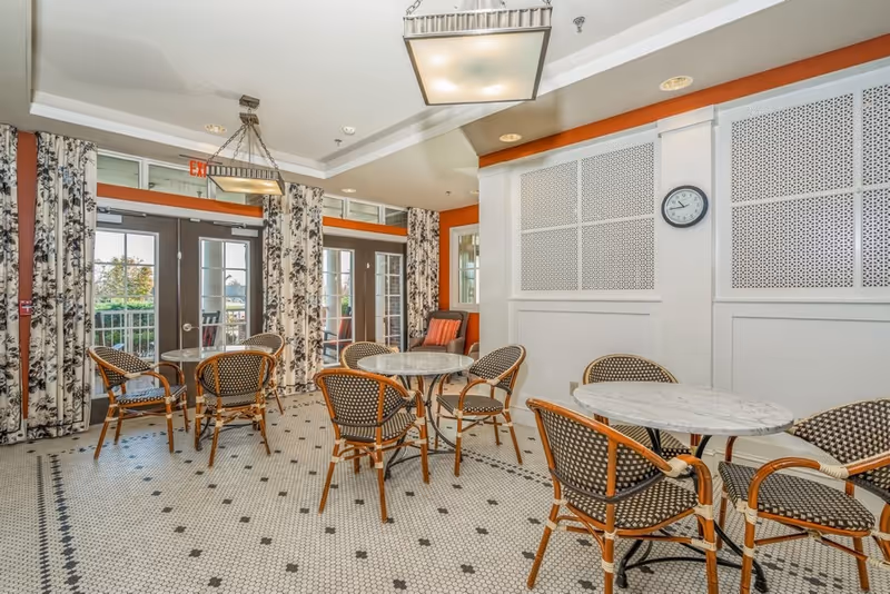 A bright dining area with round marble-top tables and wicker chairs arranged on a patterned tile floor. The room features large windows and glass doors with floral curtains, allowing natural light to enter. A wall clock is mounted on a white paneled wall, and modern square ceiling lights hang above the tables.