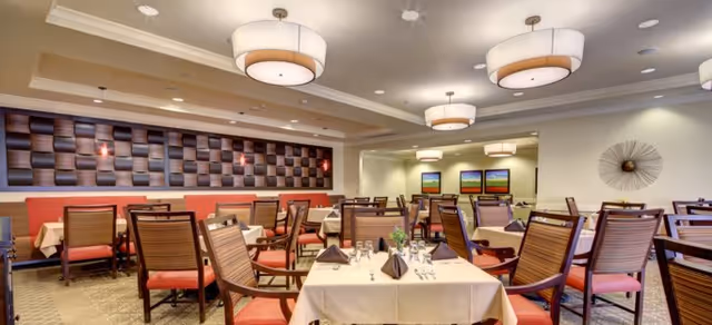 A spacious dining room with multiple tables covered in white tablecloths, set with napkins, glasses, and silverware. The room features wooden chairs with red cushions, modern ceiling lights, and decorative wall art including a textured panel and a circular metal sculpture.