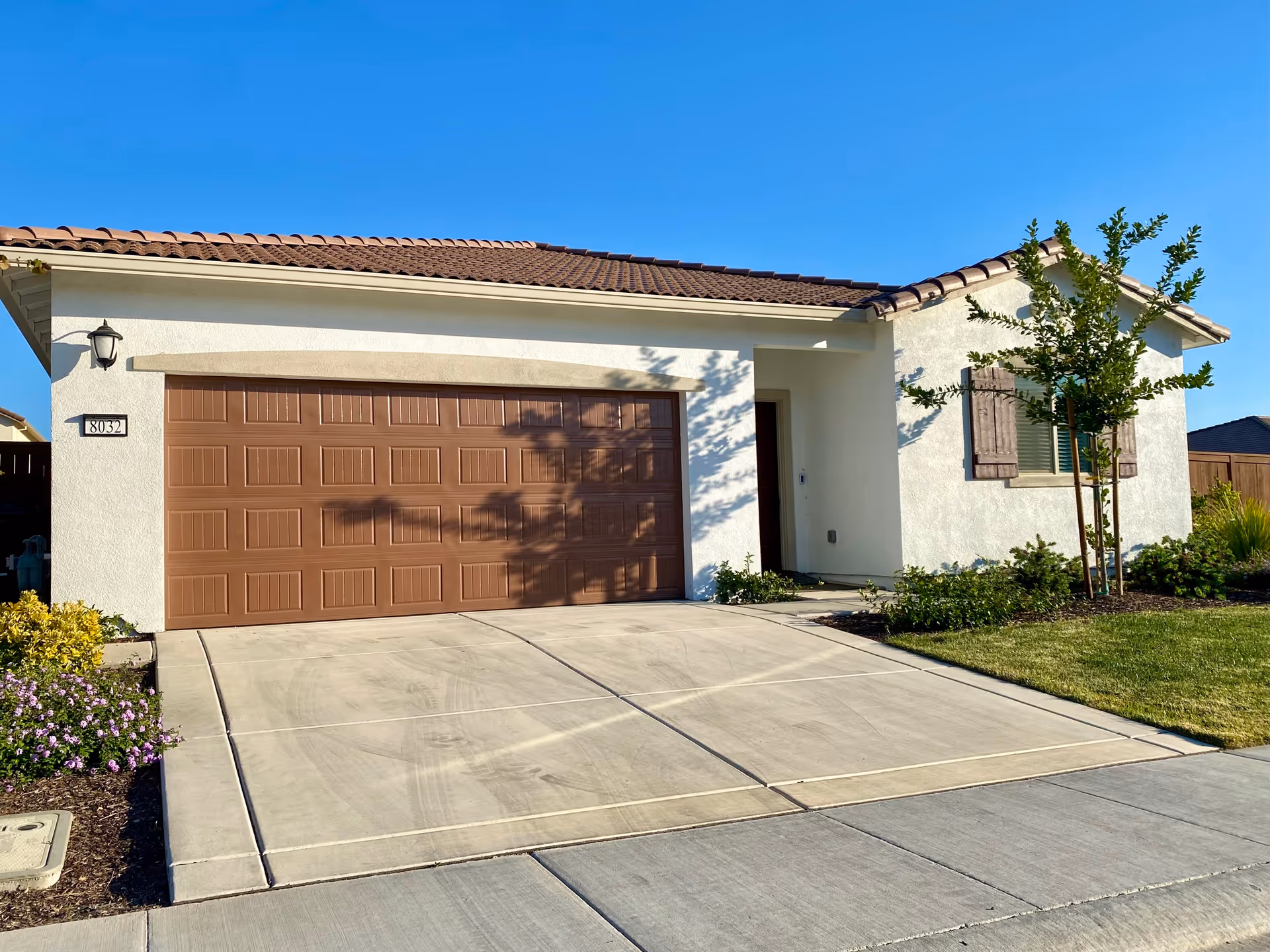 Front exterior of a single-story house with a closed brown garage door, concrete driveway, and small landscaped yard under a clear blue sky.