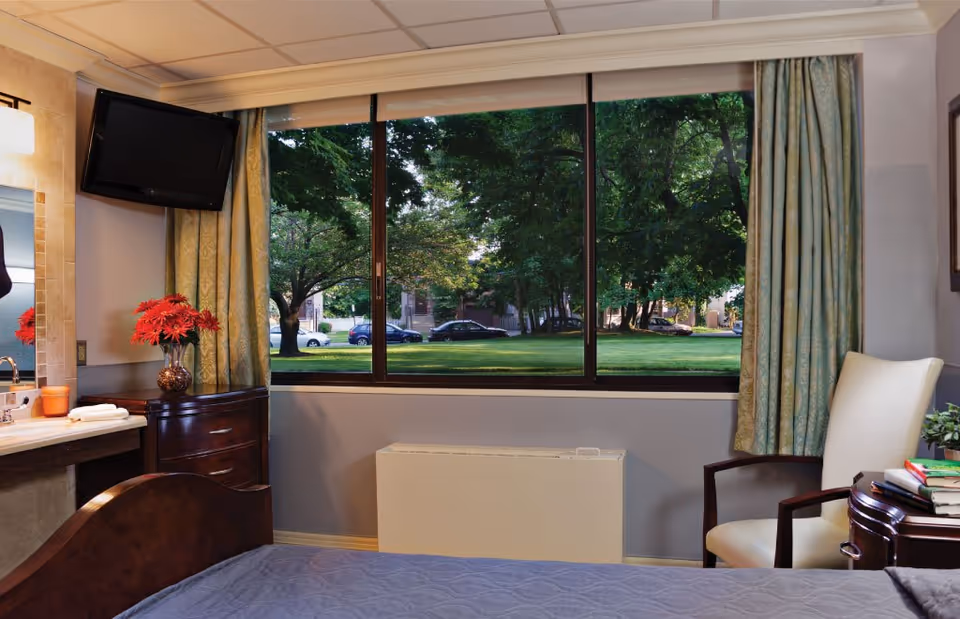 A cozy bedroom in a senior living facility with a large window showing a green outdoor area with trees and parked cars. The room features a bed with a blue quilt, a wooden nightstand with a vase of red flowers, a wall-mounted TV, a white armchair, and a small table with books and a plant.