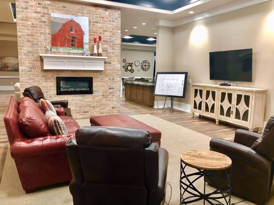 A cozy living room area with a red leather sofa, two dark leather armchairs, and a matching red ottoman. The room features a brick fireplace with a white mantel, decorated with a painting of a red barn and some candle holders. A flat-screen TV is mounted on the wall above a wooden cabinet. In the background, there is a kitchen island and a wall with several decorative clocks.