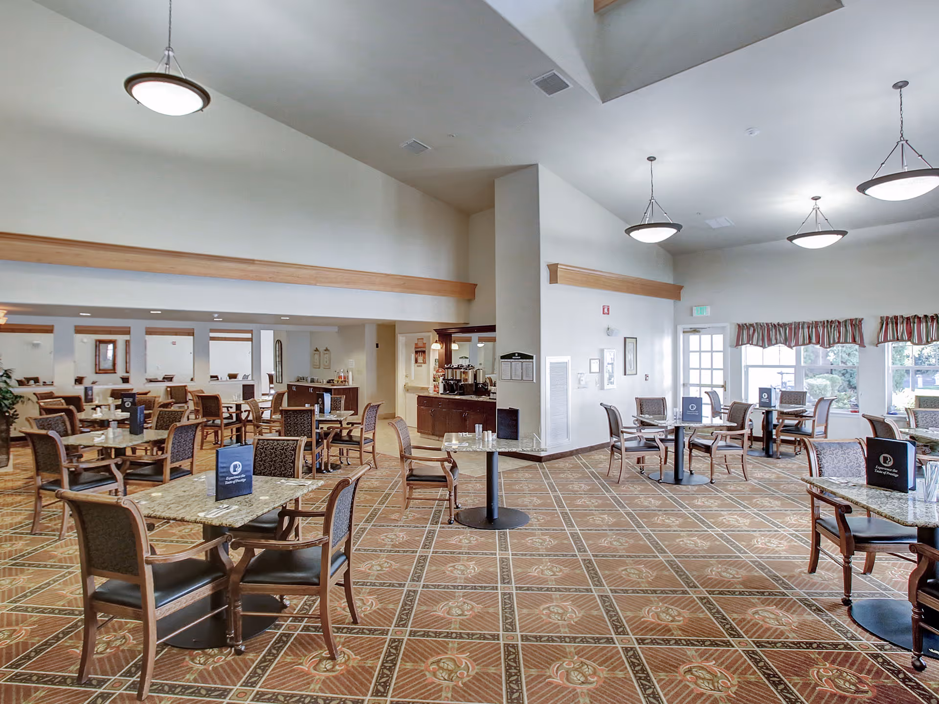 A spacious dining room with multiple tables and chairs arranged neatly. The room features patterned carpet flooring, high ceilings with hanging light fixtures, and large windows with valance curtains allowing natural light to enter. There is a small counter area with coffee machines and a doorway leading to another room.