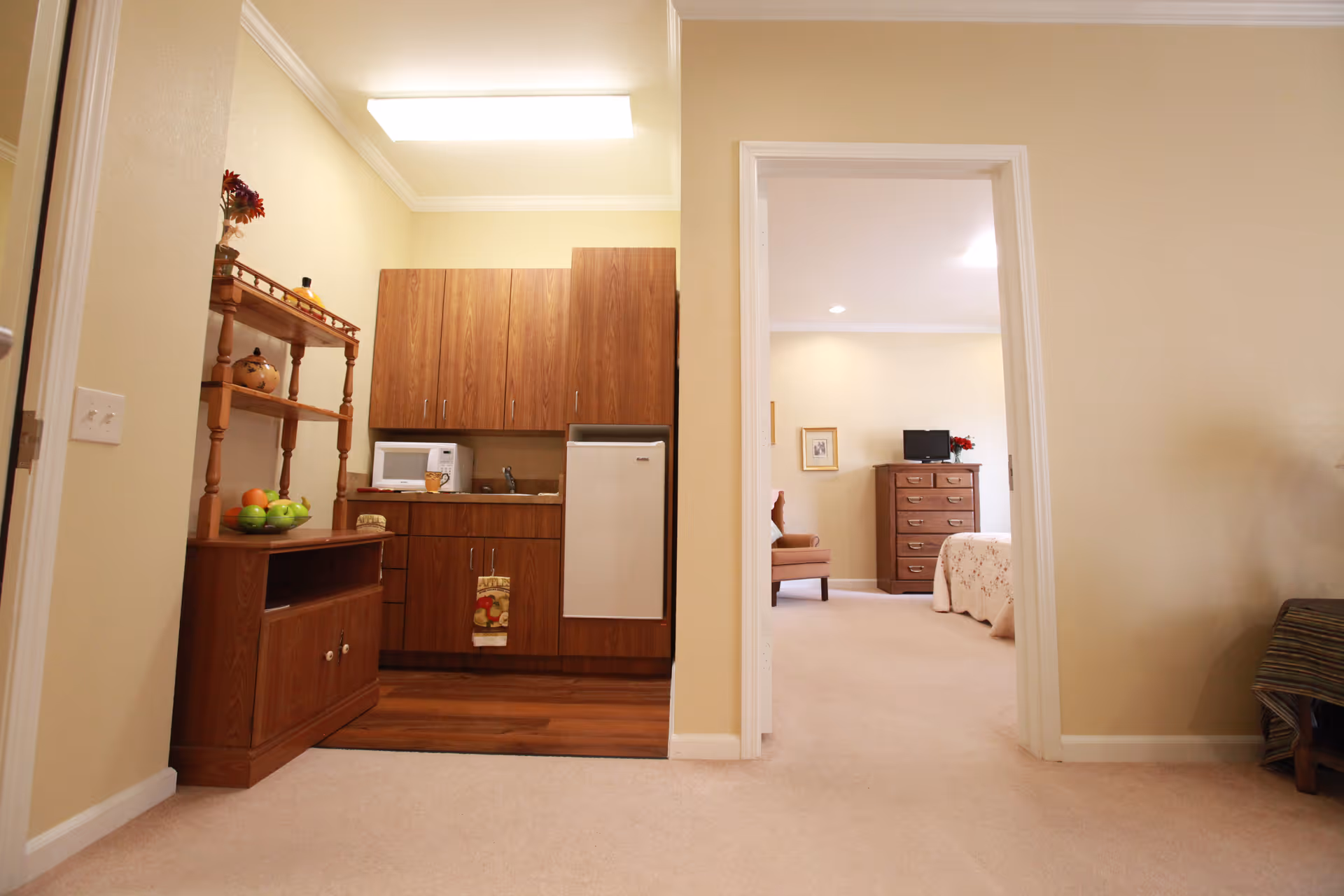 Interior view of a senior living facility showing a small kitchenette area with wooden cabinets, a microwave, a mini fridge, and a wooden shelf with decorative items and fruit. To the right, an open doorway leads to a bedroom with a bed, a dresser with a TV on top, and a chair. The walls are painted light beige and the floor is carpeted.