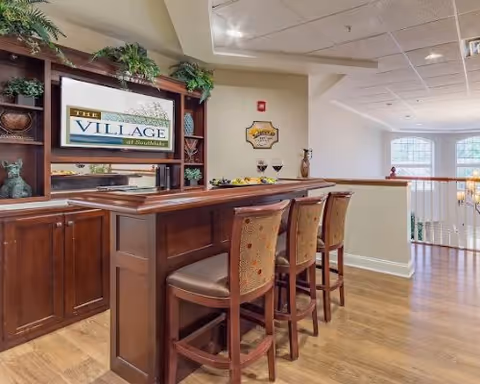 Interior view of a senior living facility lounge area featuring a wooden bar counter with four high chairs. Behind the bar is a wooden shelving unit with decorative plants and a sign that reads 'The Village at Southlake.' The room has light-colored walls, large windows, and wooden flooring.