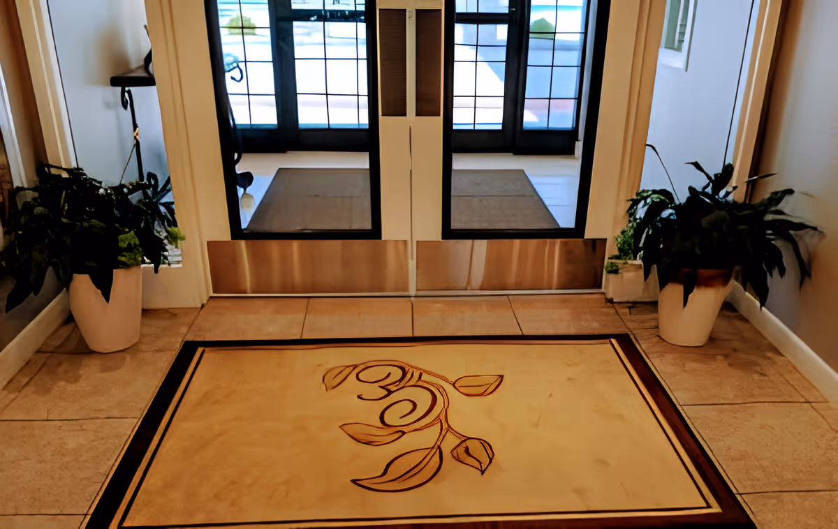 Entrance area of Courtyard Estates of Knoxville featuring double glass doors with black frames, a beige floor mat with a decorative leaf design, tiled flooring, and potted plants on either side of the doors.