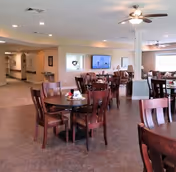 A spacious dining area with several round wooden tables and matching chairs. The room has a ceiling fan with lights, large windows letting in natural light, and a television mounted on the far wall. The walls are painted a light color, and there is a hallway visible to the left.