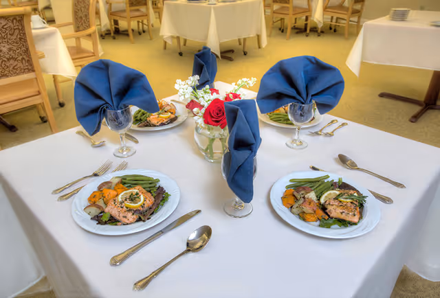 A dining table set for three with white tablecloth, blue folded napkins in glasses, and plates of cooked salmon with vegetables. A small vase with red and white flowers is in the center. The background shows more tables and chairs in a dining room setting.