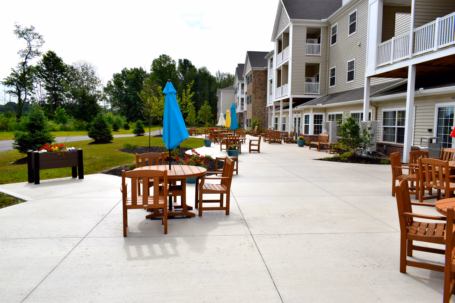 Outdoor patio area at Danbury Hudson with multiple wooden tables and chairs, some tables have closed blue and yellow umbrellas. The patio is adjacent to a multi-story building with balconies and surrounded by greenery and trees.