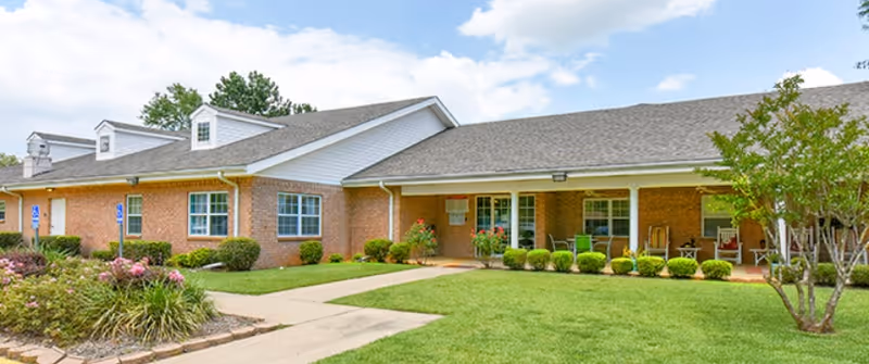 Exterior view of a single-story brick building with a covered porch, green lawn, bushes, and a tree under a partly cloudy sky.