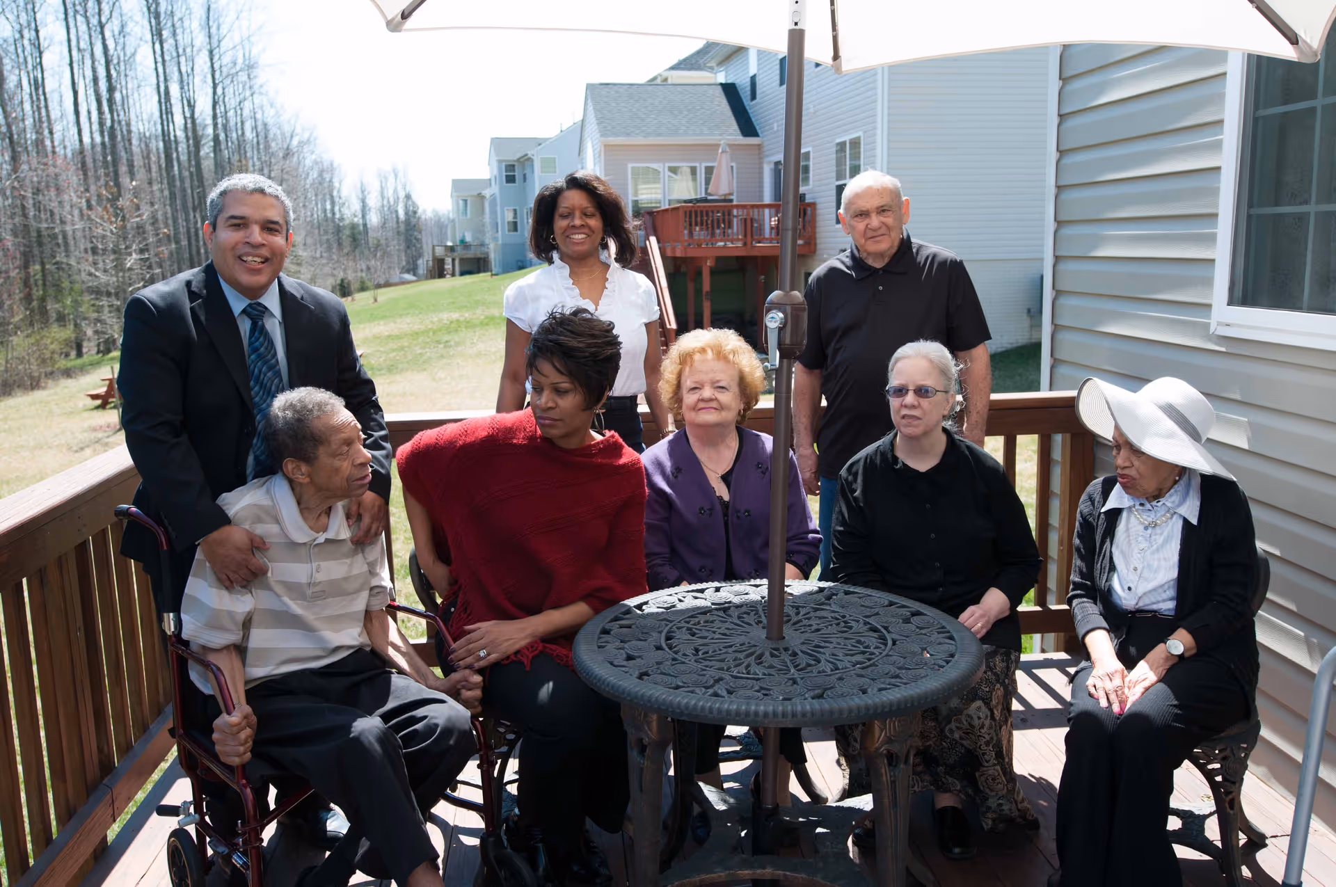 A group of seven people, including elderly individuals and caregivers, gathered on a wooden outdoor deck with a round metal table and umbrella stand in the center. The background shows a grassy area and neighboring houses under a clear sky.