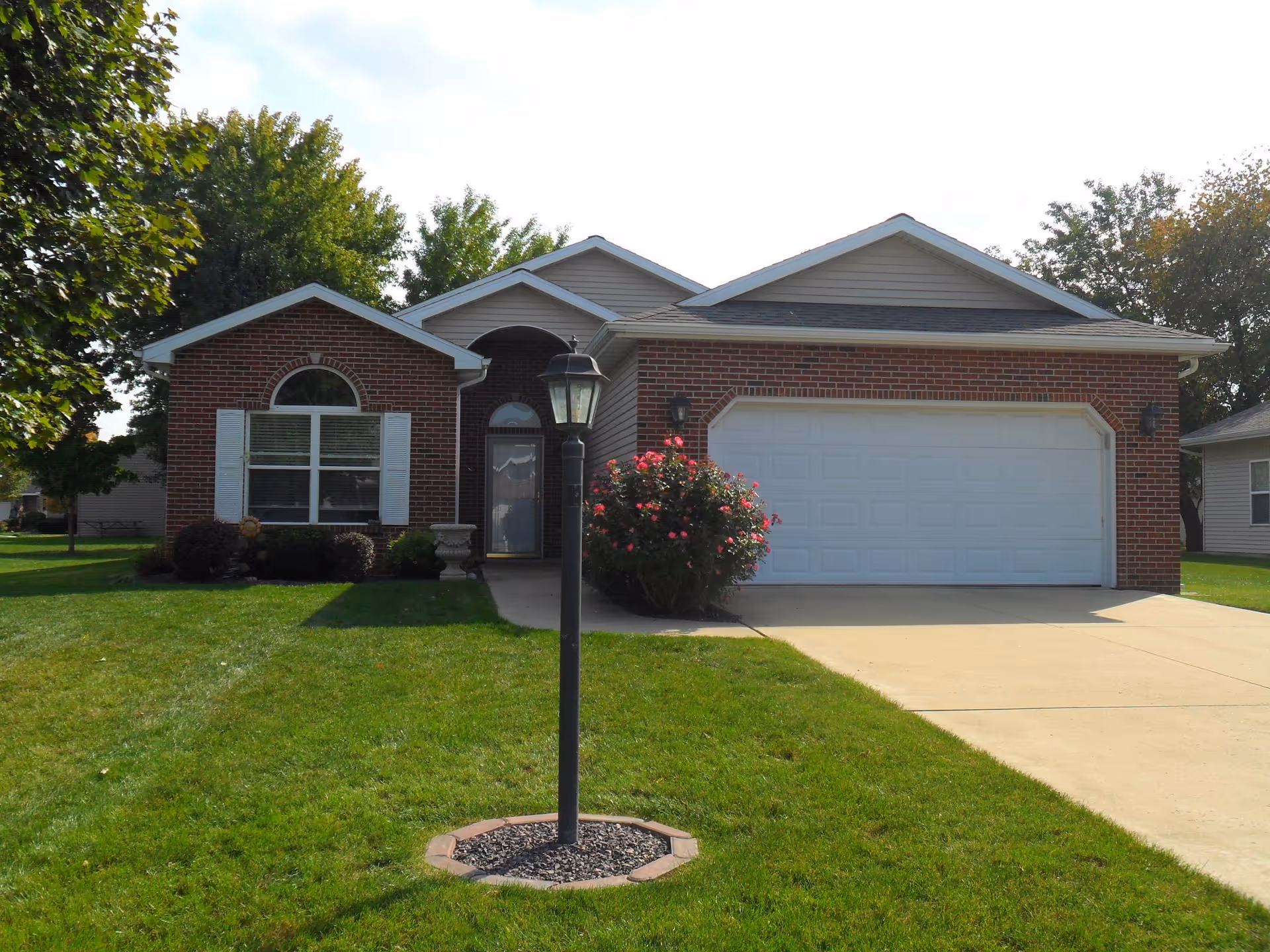 Front exterior view of a single-story brick house with a white garage door, a concrete driveway, a lamp post in the front yard, green grass, and trees in the background.