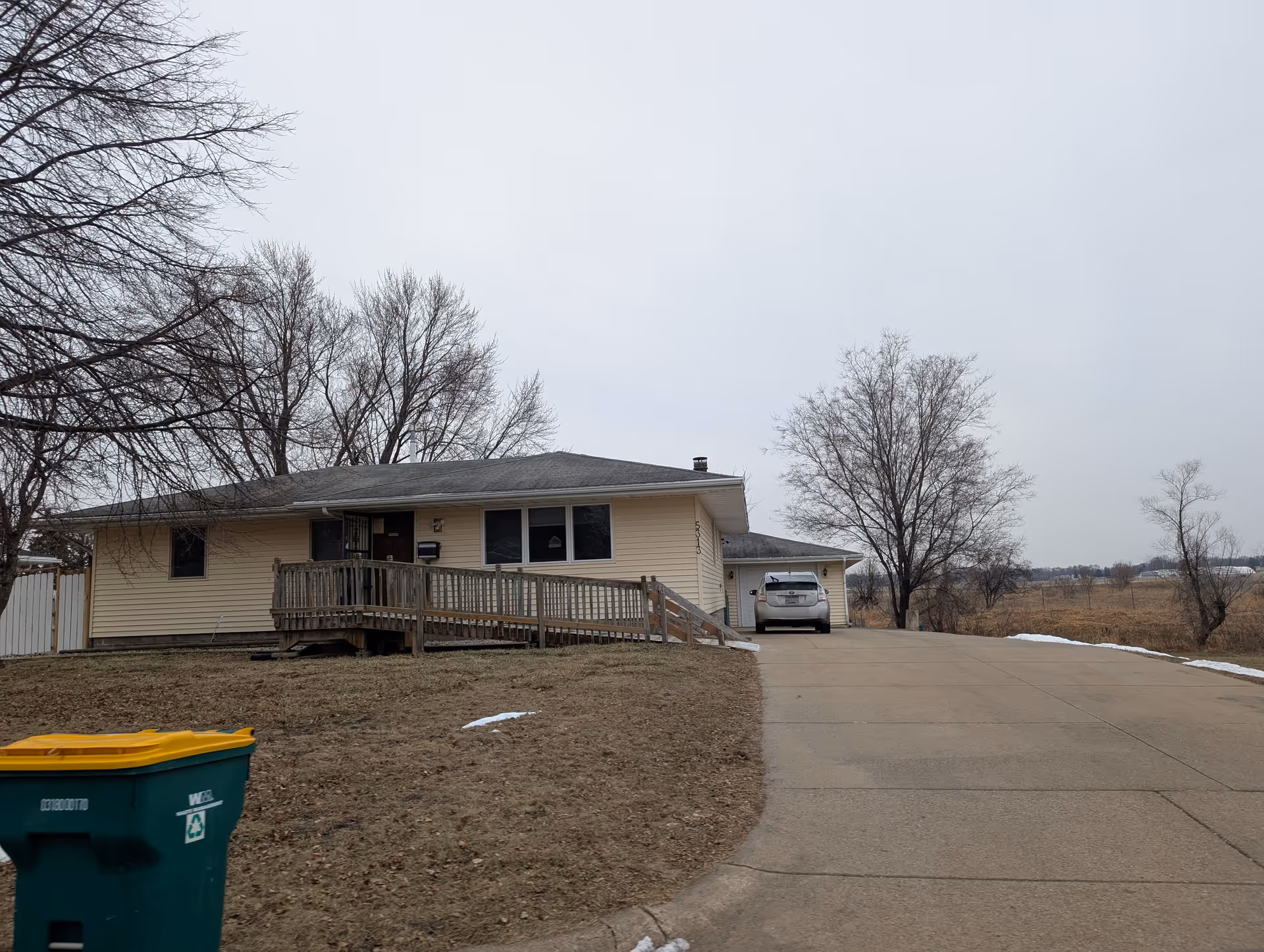 Single-story beige house with a wheelchair ramp leading to the front door, a driveway with a parked car, leafless trees, and a recycling bin in the foreground on a cloudy day.