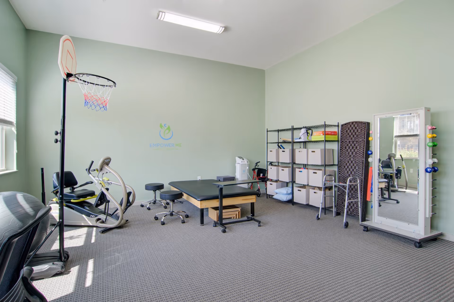 A therapy or exercise room with light green walls and carpeted floor. The room contains a basketball hoop, exercise bike, therapy table, stools, storage shelves with boxes, a walker, a large mirror with colorful hand weights attached, and a window letting in natural light.