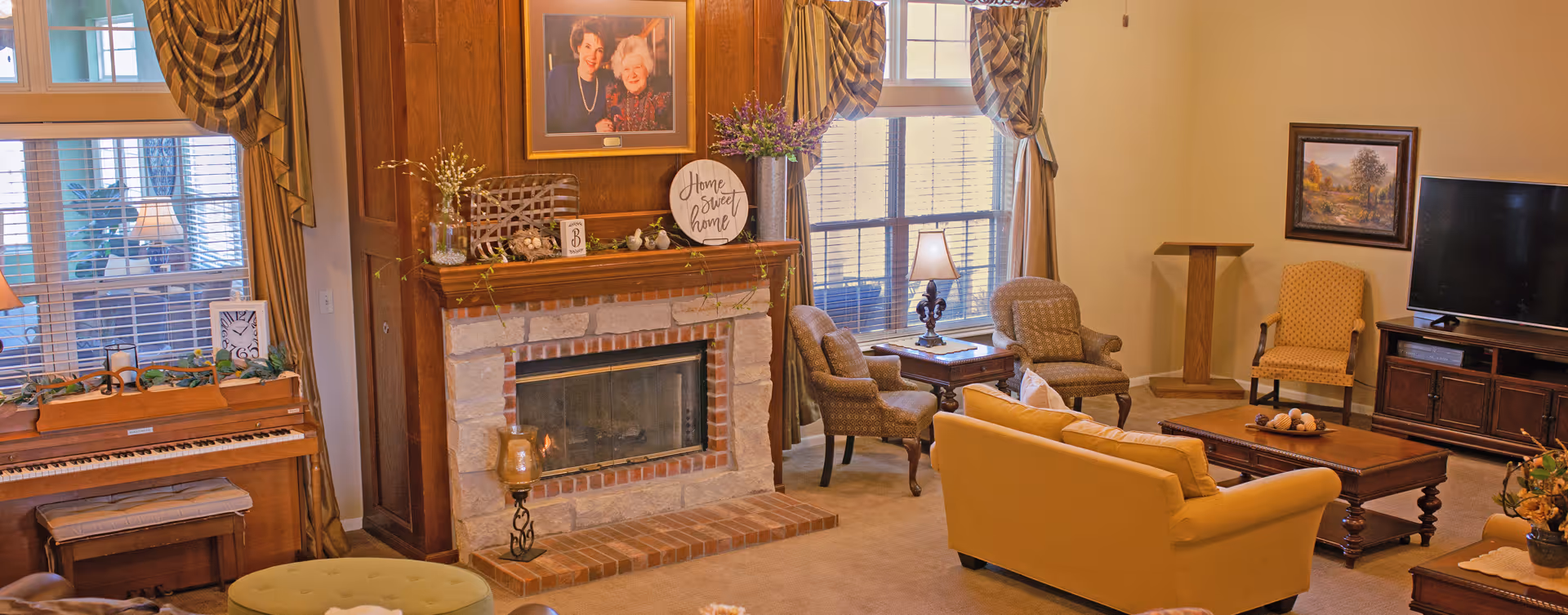 A cozy living room with a stone and brick fireplace, a wooden piano with a bench, several upholstered chairs, a yellow sofa, a wooden coffee table, a TV on a wooden stand, and framed pictures on the walls. The room has large windows with curtains and a warm, inviting atmosphere.