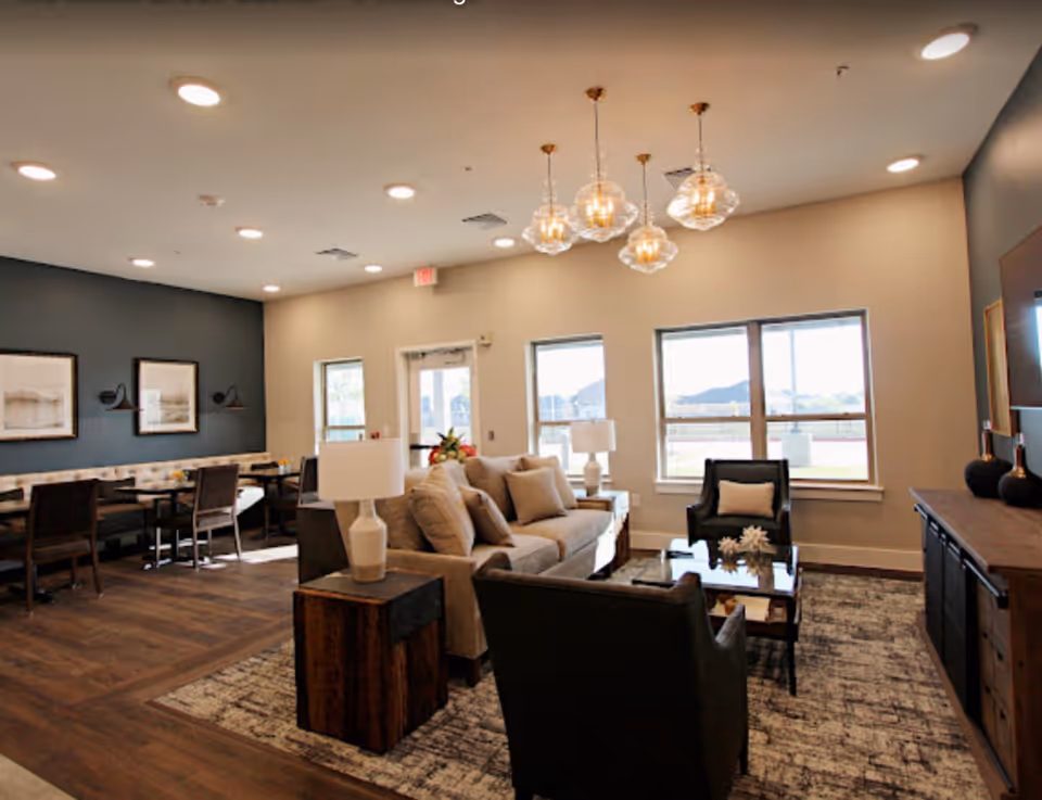 Bright, modern communal living room with sofas, armchairs, a coffee table and a dining area under pendant lights.