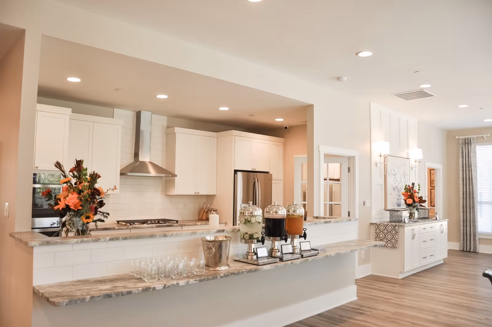 A bright, modern kitchen area with white cabinets, a stainless steel refrigerator, and a stove with a stainless steel hood. The kitchen counter has three beverage dispensers with different drinks, a metal ice bucket, and several empty glasses. There are two flower arrangements, one on the kitchen counter and one on a sideboard against the wall. The room has recessed lighting and wood flooring.