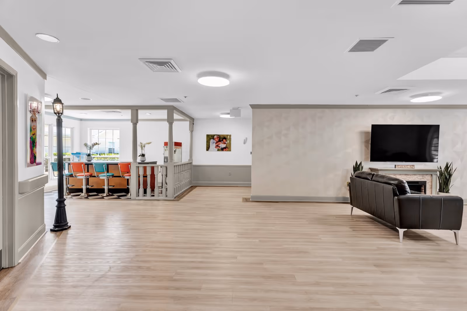 Spacious interior common area with light wood flooring, a black leather sofa facing a wall-mounted flat screen TV above a fireplace, and a dining area with colorful chairs and a popcorn machine in the background. The room is well-lit with ceiling lights and has decorative plants and artwork on the walls.